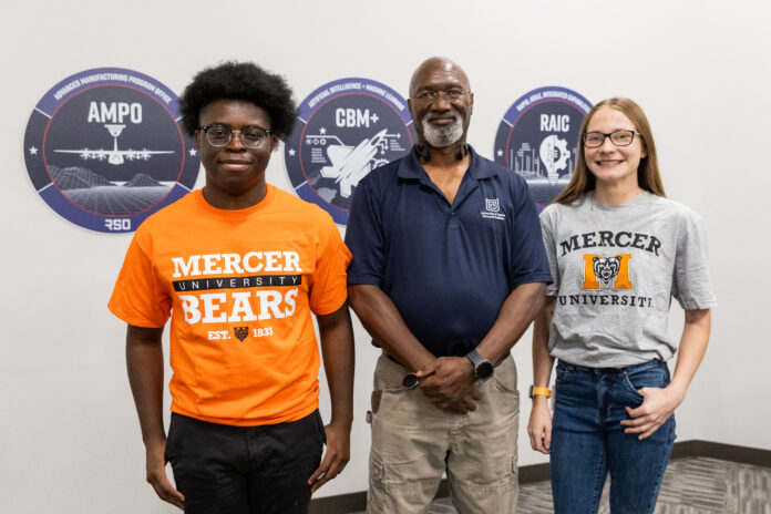 Three people stand indoors in front of wall signs, two wearing Mercer University shirts and one in a navy polo.