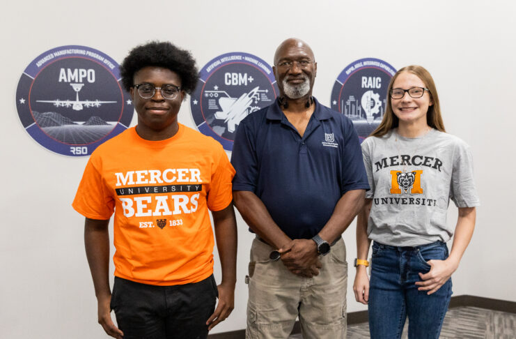 Three people stand indoors in front of wall signs, two wearing Mercer University shirts and one in a navy polo.