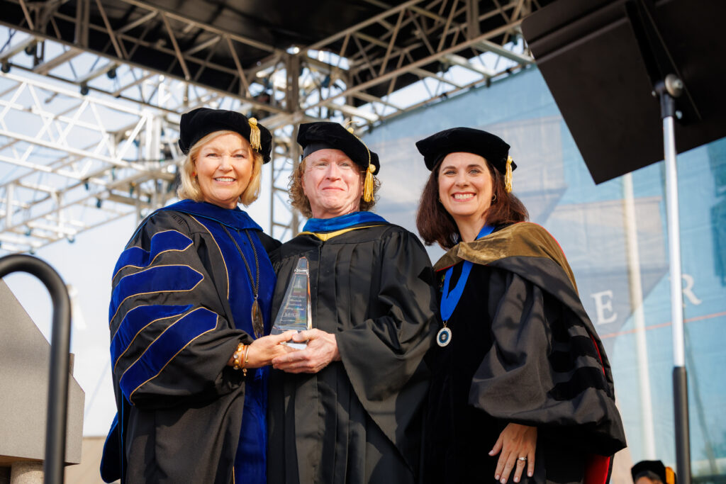 Three people in academic regalia stand together at a graduation ceremony; one holds a glass award.