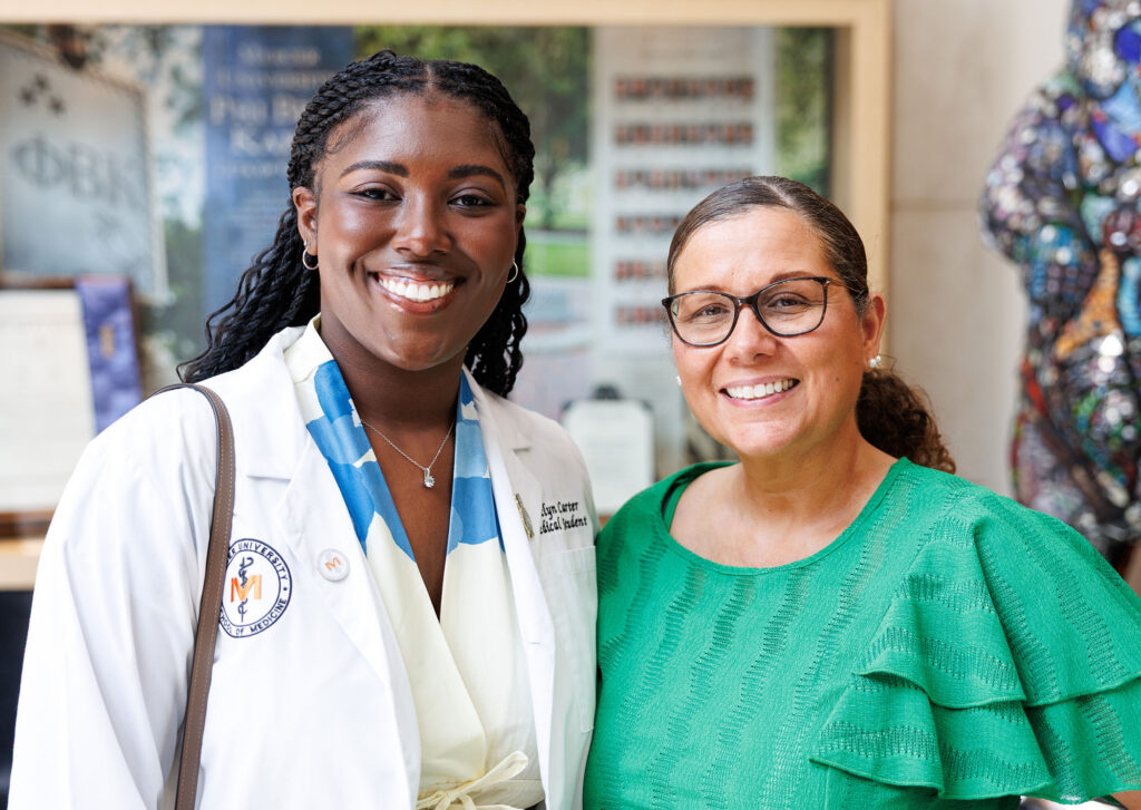 Two women smiling, one in a white medical coat and the other in a green top, standing indoors.