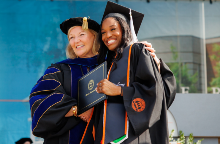 A graduate and faculty member in academic regalia smile and pose together at a graduation ceremony.