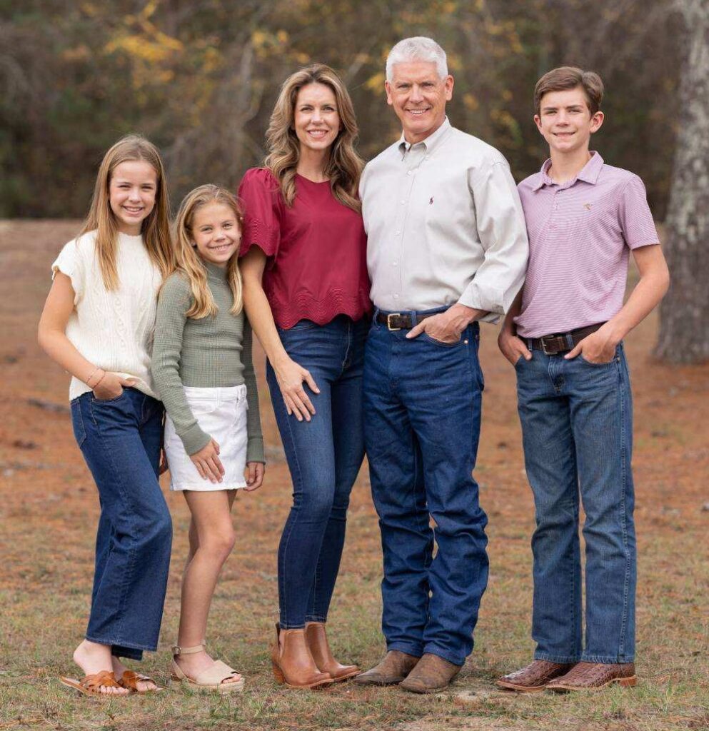 Five people, two adults and three children, standing outdoors on grass, posing for a family photo, all smiling at the camera.