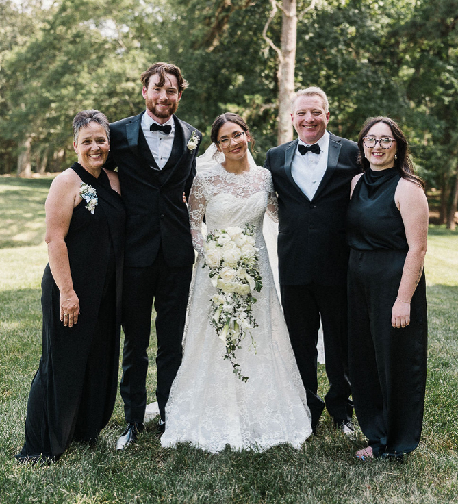 Five people pose together outdoors, including a bride in a white dress holding a bouquet and four others in formal black attire.