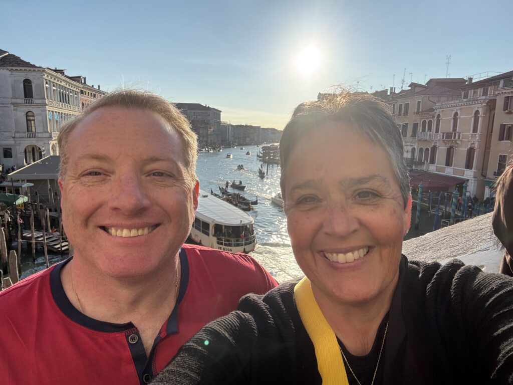 Two people smiling for a selfie with a canal, buildings, and boats in the background during sunset.