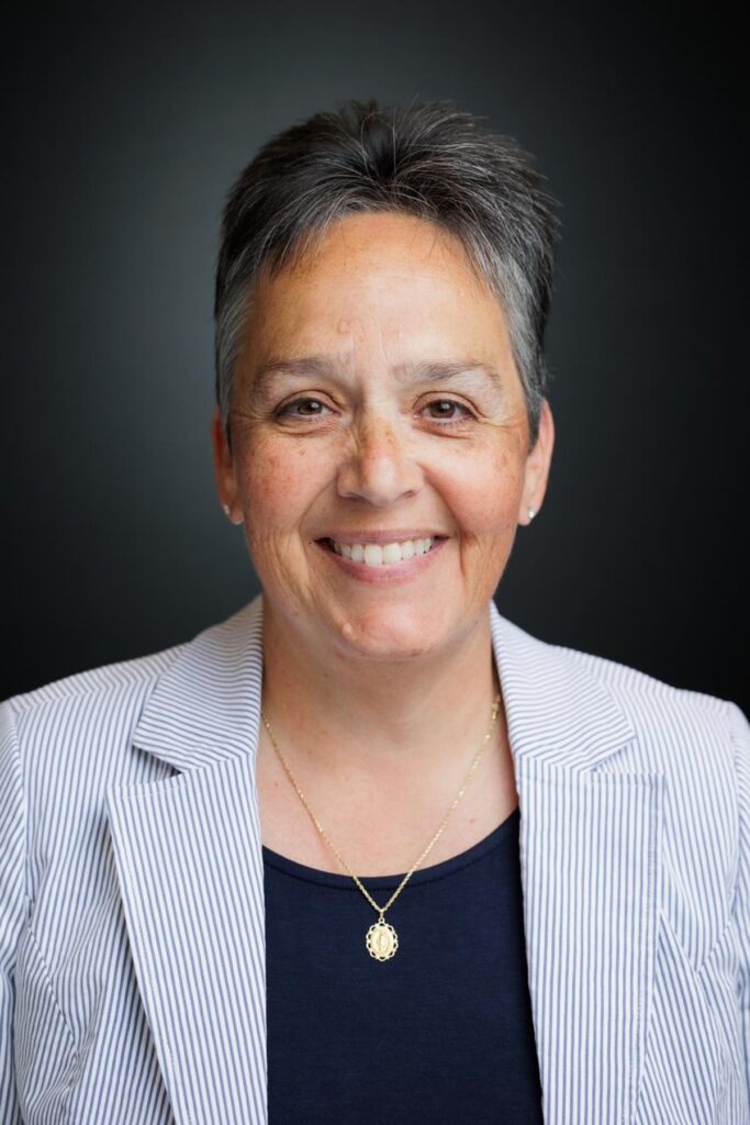 Smiling woman with short gray hair wearing a light striped blazer and navy shirt, posing against a dark background.