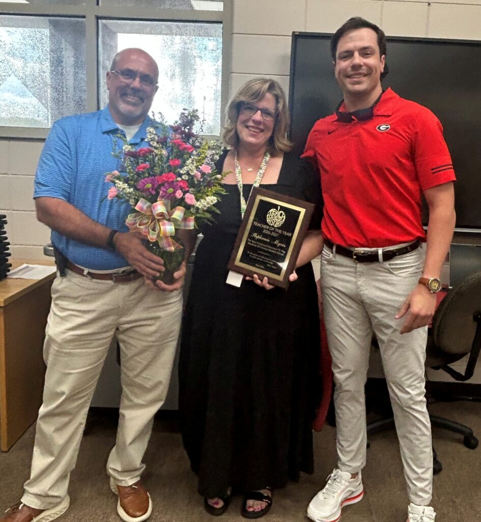 Three people stand indoors; one holds flowers, another holds a plaque, and they are all smiling at the camera.