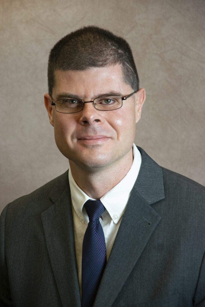 Man wearing glasses, a dark suit, white shirt, and navy tie, posed in front of a neutral background.