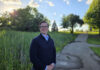 Alumnus works to develop new cancer treatments A person stands smiling on a paved path in a park with trees and tall grass under a sunny sky.