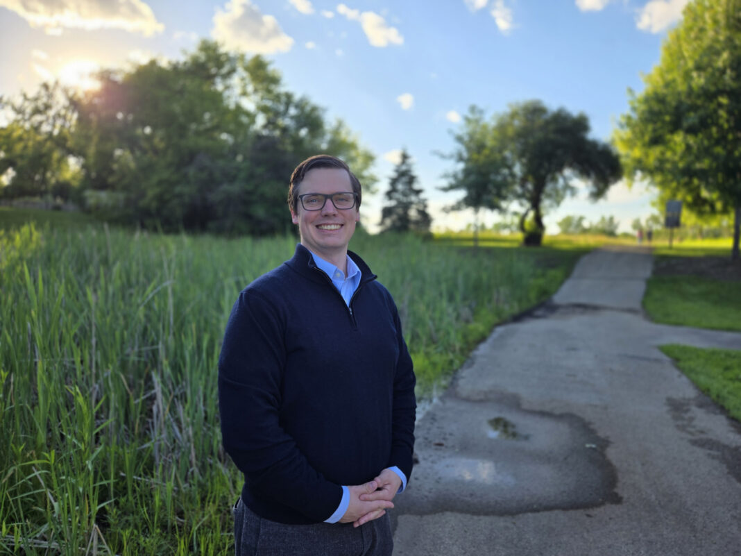 A person stands smiling on a paved path in a park with trees and tall grass under a sunny sky.