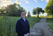 Alumnus works to develop new cancer treatments A person stands smiling on a paved path in a park with trees and tall grass under a sunny sky.