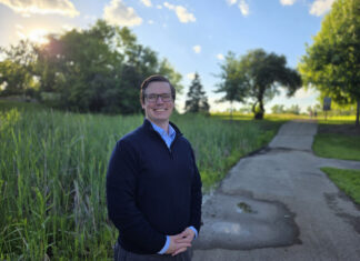 Alumnus works to develop new cancer treatments A person stands smiling on a paved path in a park with trees and tall grass under a sunny sky.