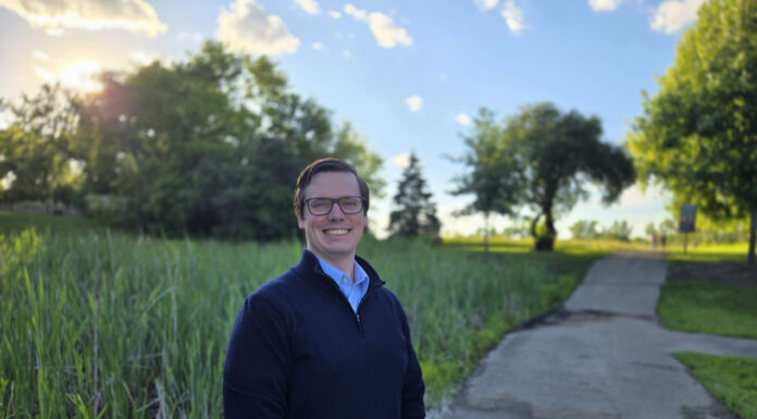 Alumnus works to develop new cancer treatments A person stands smiling on a paved path in a park with trees and tall grass under a sunny sky.