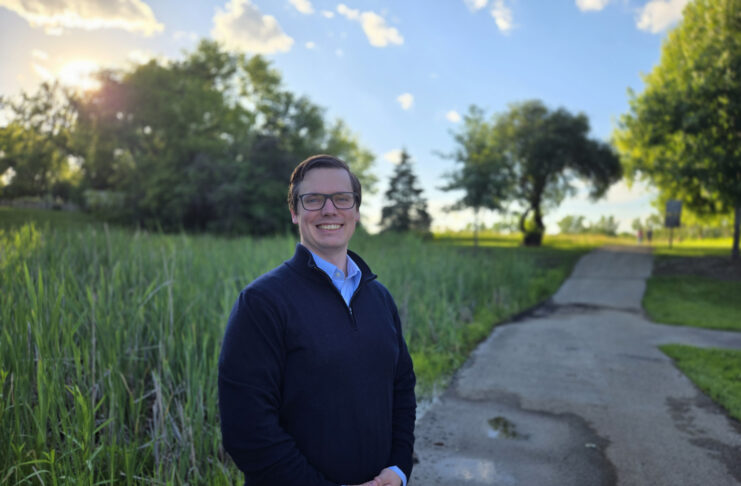 A person stands smiling on a paved path in a park with trees and tall grass under a sunny sky.