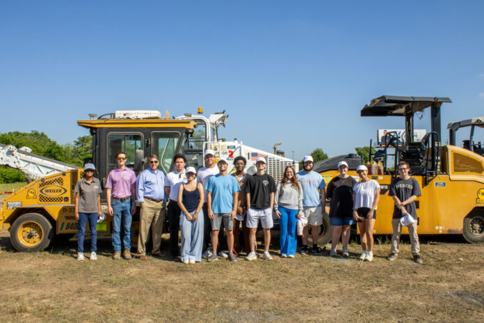 A group of people standing outdoors in front of large construction vehicles under a clear blue sky.