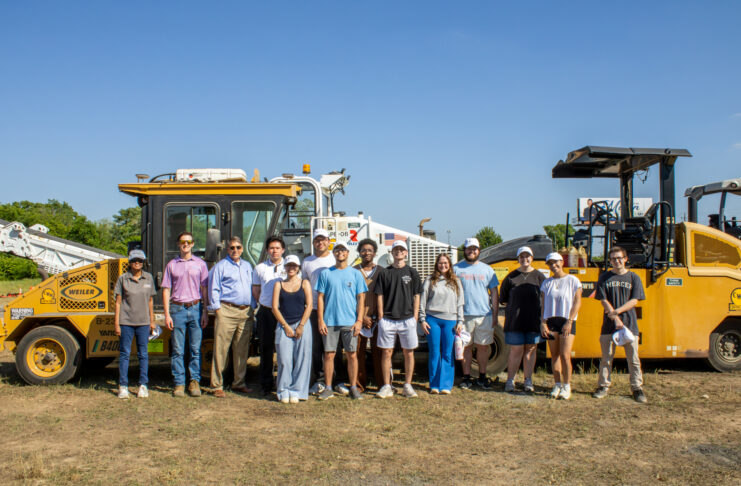 A group of people standing outdoors in front of large construction vehicles under a clear blue sky.