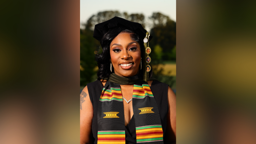 A woman in graduation attire with a kente stole smiles outdoors with trees in the background.