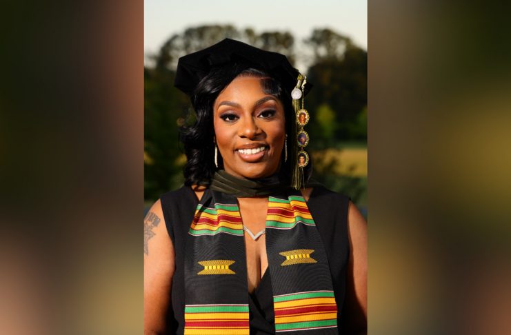 A woman in graduation attire with a kente stole smiles outdoors with trees in the background.