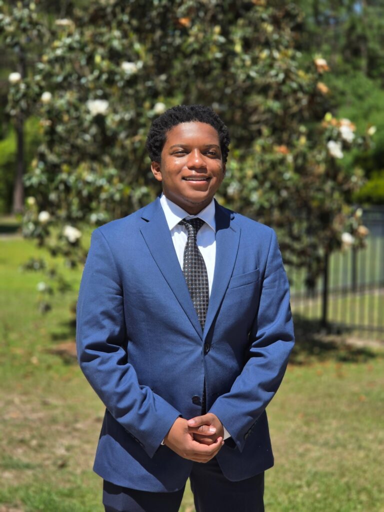 A young man wearing a blue suit and tie stands outdoors in front of a tree on a sunny day.