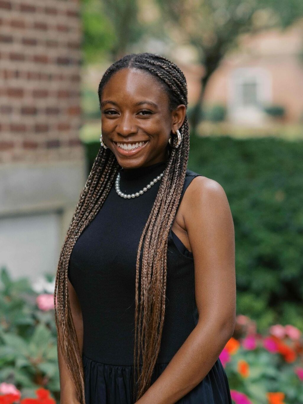 A woman with long braids and a black dress smiles while standing in a garden with flowers and greenery.