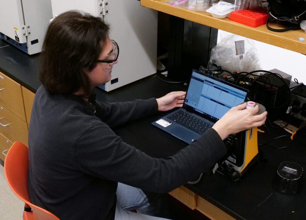 A person in safety glasses operates lab equipment and monitors data on a laptop at a laboratory workstation.
