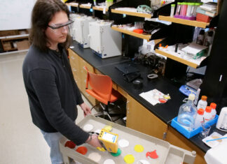 Mercer professors develop international standard for detecting lead in new paint Person in safety glasses uses a handheld device to scan paint samples on a cart in a laboratory setting.