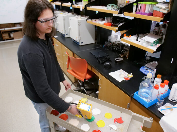 Person in safety glasses uses a handheld device to scan paint samples on a cart in a laboratory setting.