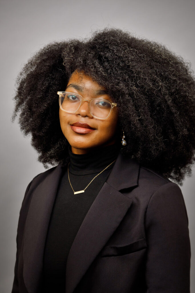 A woman with natural curly hair, glasses, and a black blazer poses against a plain gray background.