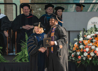 Mercer scholar-athletes post record year in the classroom Graduate in cap and gown shakes hands with faculty member on stage at a commencement ceremony with flowers nearby.
