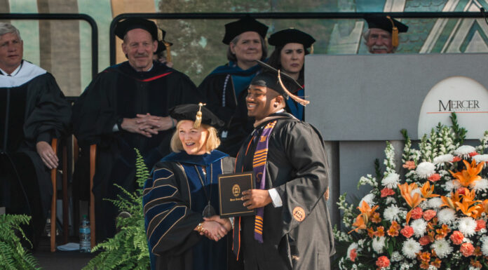 Mercer scholar-athletes post record year in the classroom Graduate in cap and gown shakes hands with faculty member on stage at a commencement ceremony with flowers nearby.