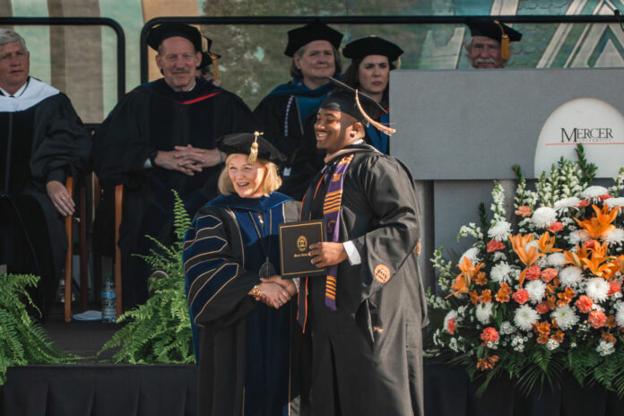 Graduate in cap and gown shakes hands with faculty member on stage at a commencement ceremony with flowers nearby.