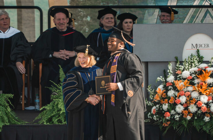 Graduate in cap and gown shakes hands with faculty member on stage at a commencement ceremony with flowers nearby.