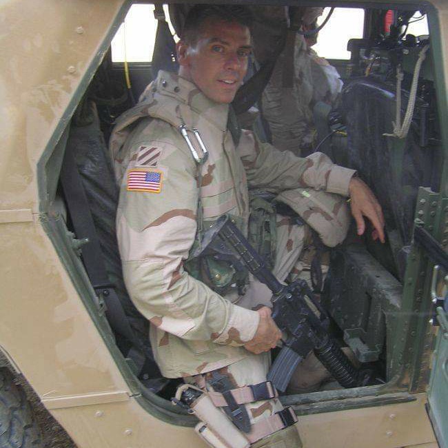 A soldier in desert camouflage gear sits inside a military vehicle holding a rifle.