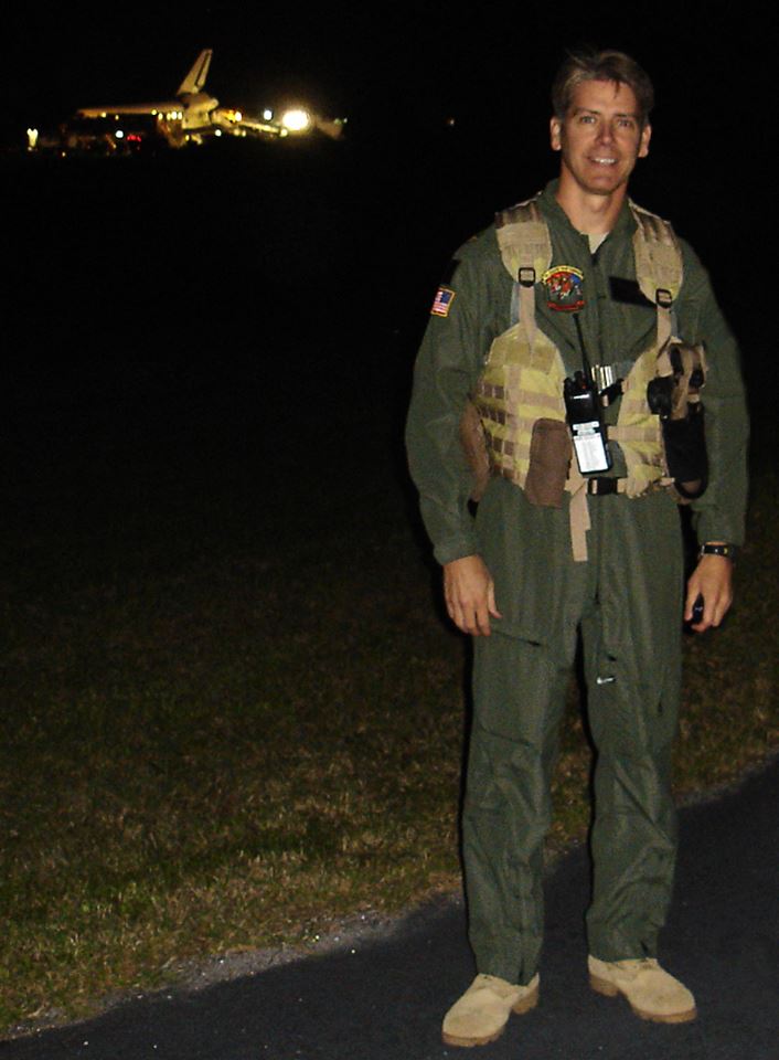 A man in a flight suit stands outdoors at night with a space shuttle visible in the background.