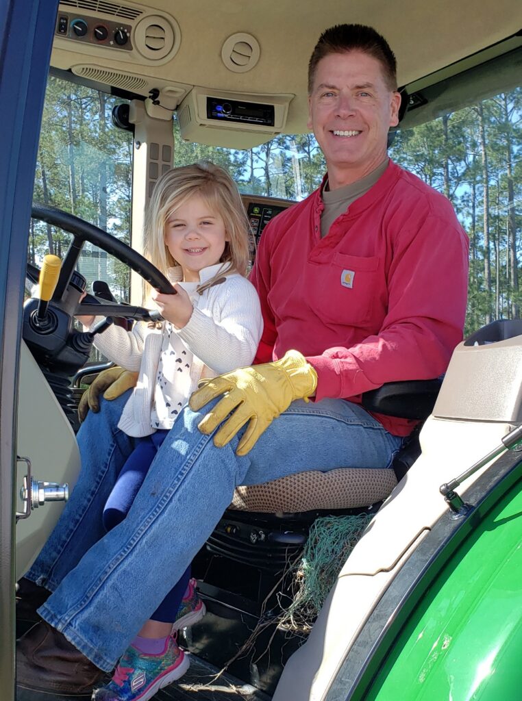Man and young girl sitting together in the cab of a green tractor, both smiling at the camera.