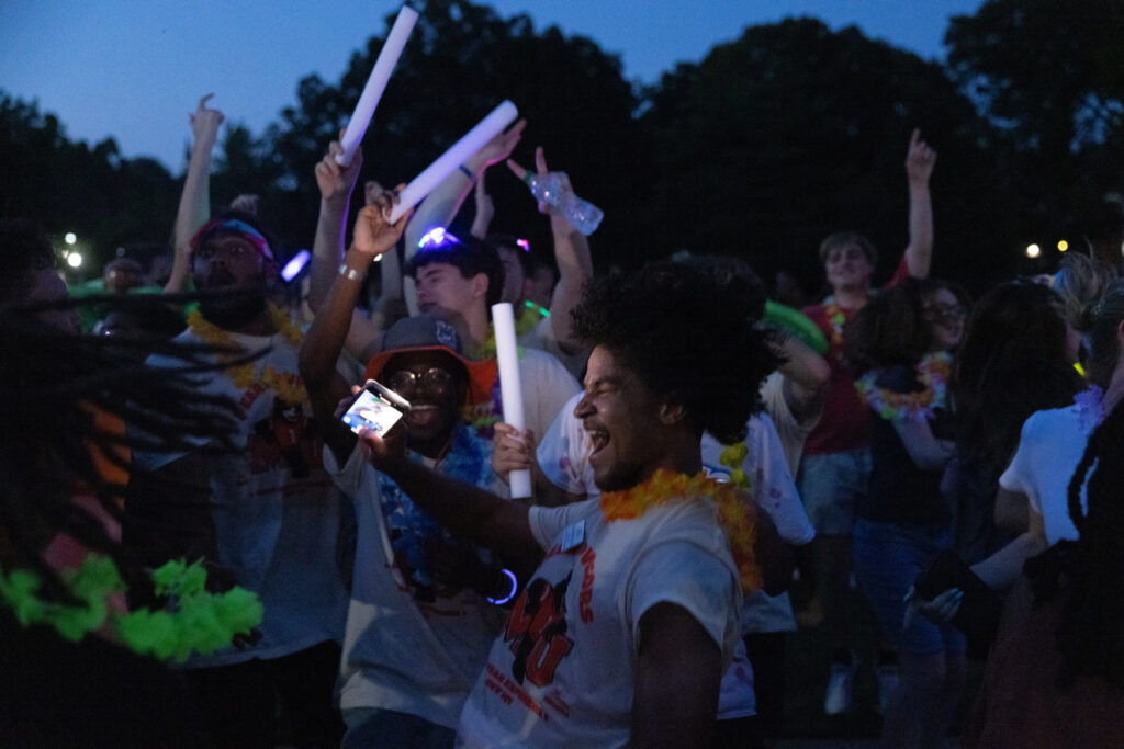 A group of people celebrate outdoors at night, wearing leis and holding glow sticks, smiling, dancing, and taking photos.