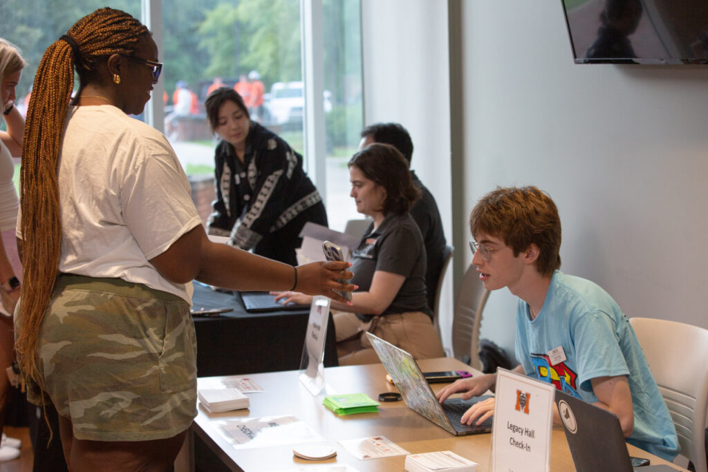A student hands her phone to a clerk at a check-in table while others assist people in the background.