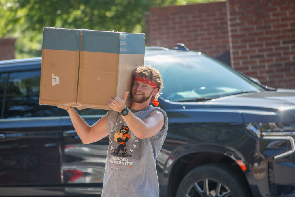 Young man in a sleeveless shirt and bandana carries a large box outdoors near a black SUV and brick wall.