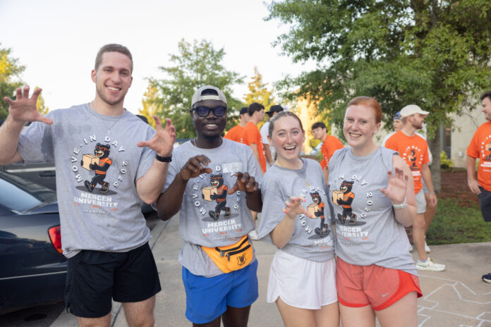 Four students in Mercer University shirts pose and smile outdoors during move-in day, with others in the background.