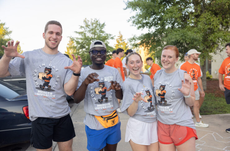 Four students in Mercer University shirts pose and smile outdoors during move-in day, with others in the background.