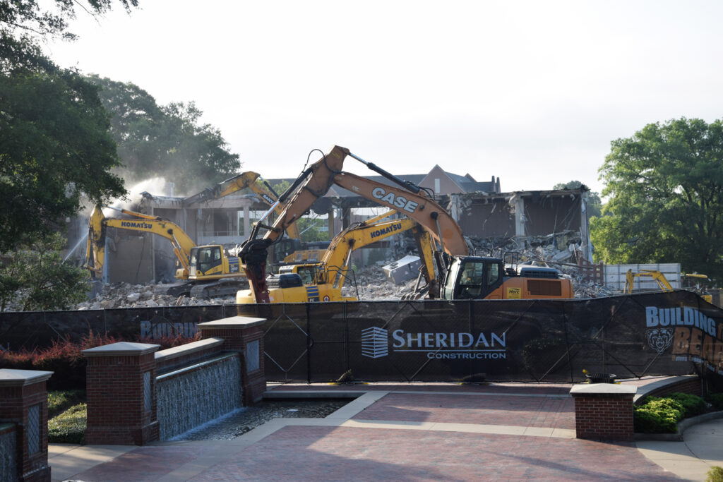Excavators demolish a large building behind a construction fence with Sheridan Construction signage.