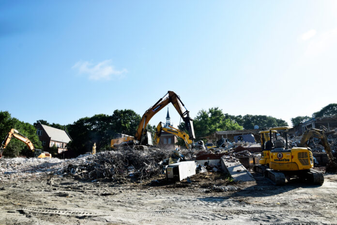 2026ConnellJanCrockerDSC0300 Excavators clearing debris and rubble at a building demolition site under a clear blue sky.