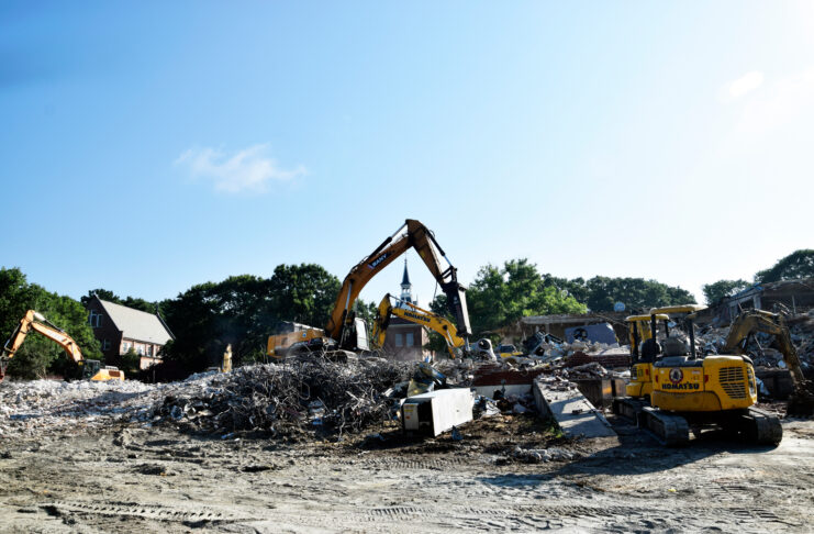 Excavators clearing debris and rubble at a building demolition site under a clear blue sky.
