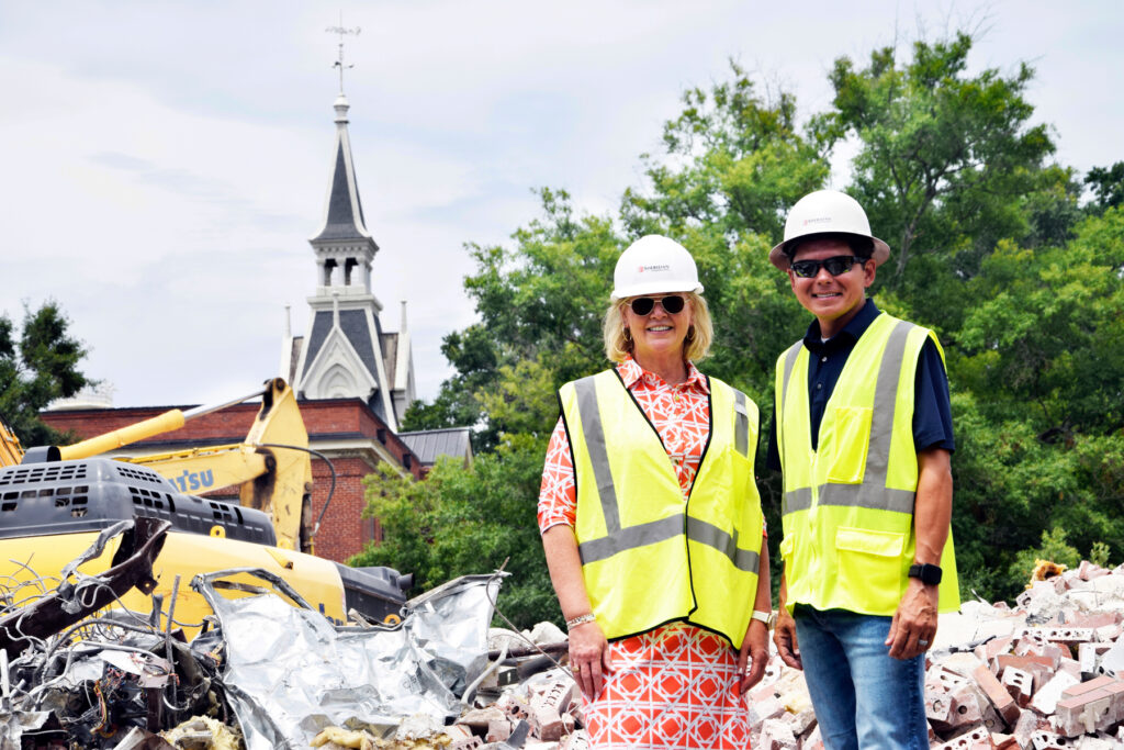 Two people in safety vests and hard hats stand at a construction site with debris, machinery, and a church in the background.