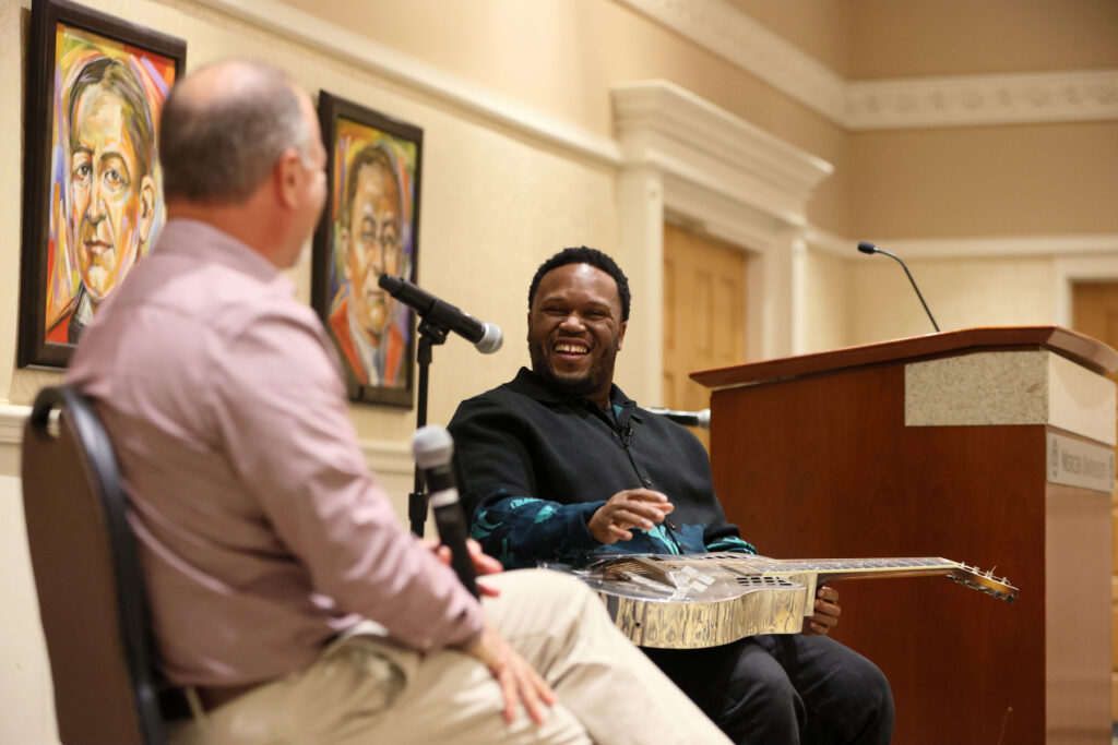 Two men sit and talk on stage with microphones; one holds a steel guitar and smiles. Framed portraits hang on the wall.
