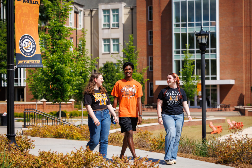 Three students walk and talk on a college campus sidewalk near a Be the Bear banner and brick buildings.