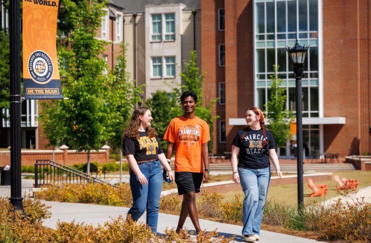 Three students walk and talk on a college campus sidewalk near a Be the Bear banner and brick buildings.