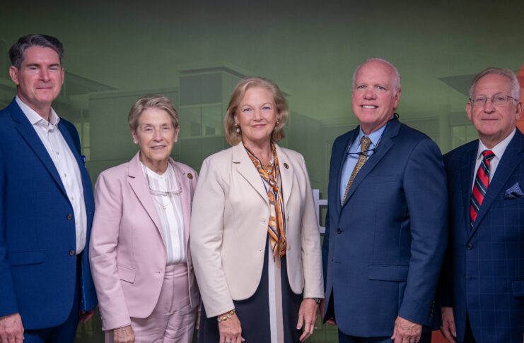 Five professionally dressed adults stand side by side against a green and red backdrop, facing the camera.