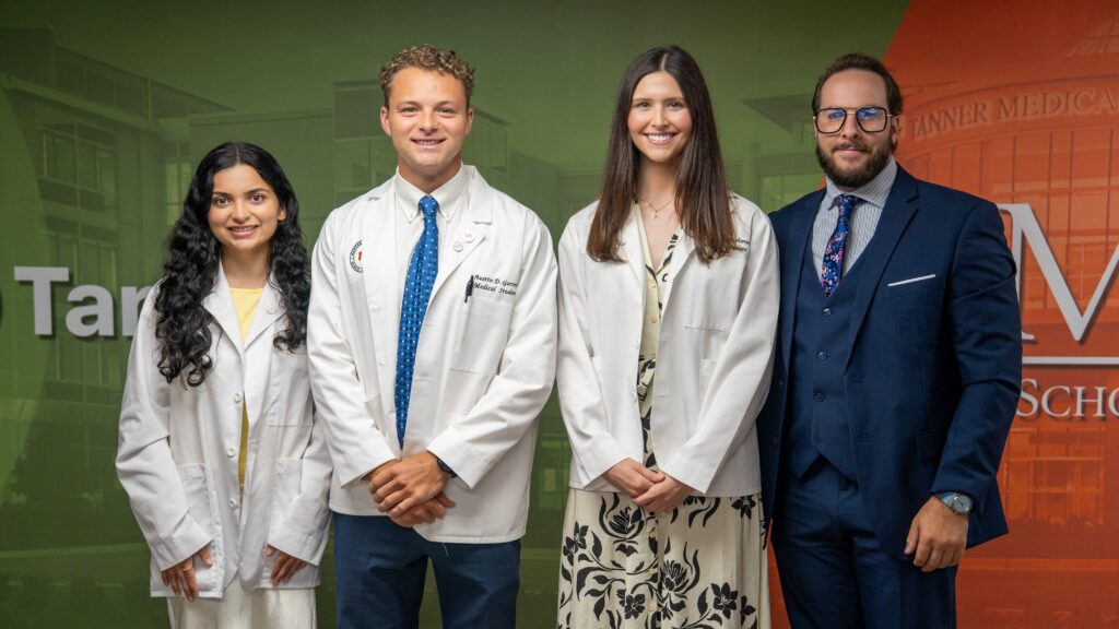 Four people, three in white medical coats and one in a suit, stand together in front of a medical school backdrop.