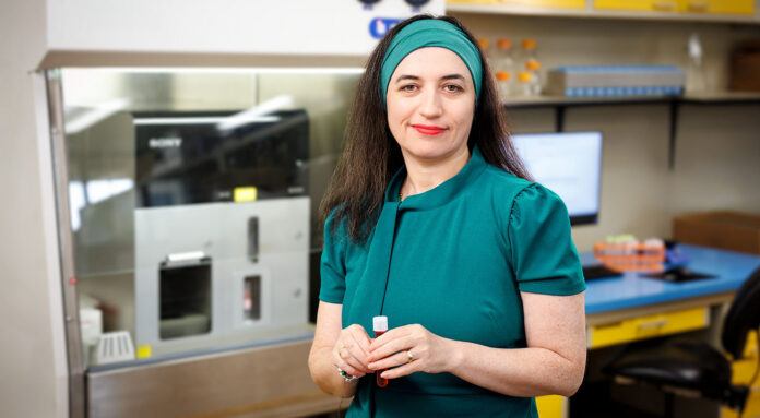 Dr. Chefetz Menaker in teal dress holding a small tube while standing in the laboratory where her research will take place.