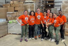 Mercer family therapy students provide behavioral healthcare to farmworkers MUSM team in matching orange shirts stand and smile in a warehouse with boxes and pallets in the background.